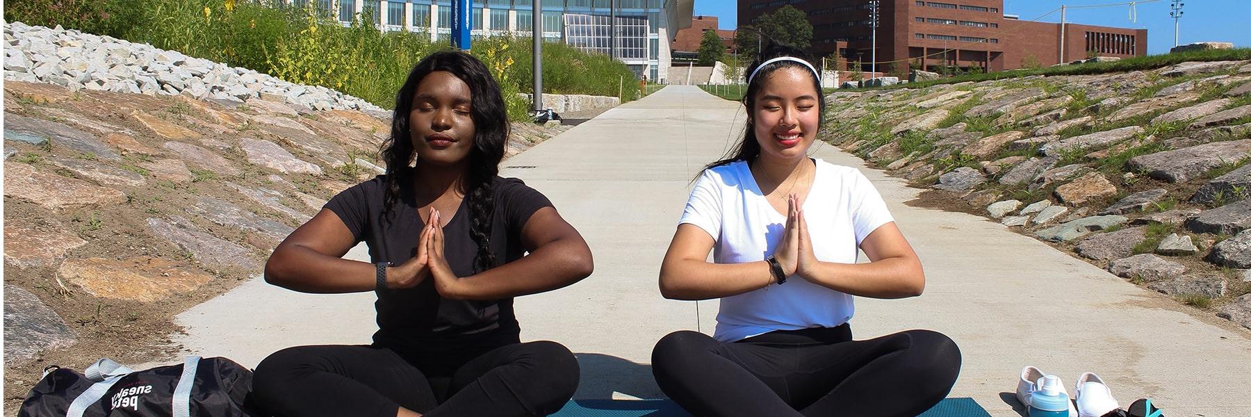 Two students sit crosslegged with hands together in a yoga pose in front of campus center.